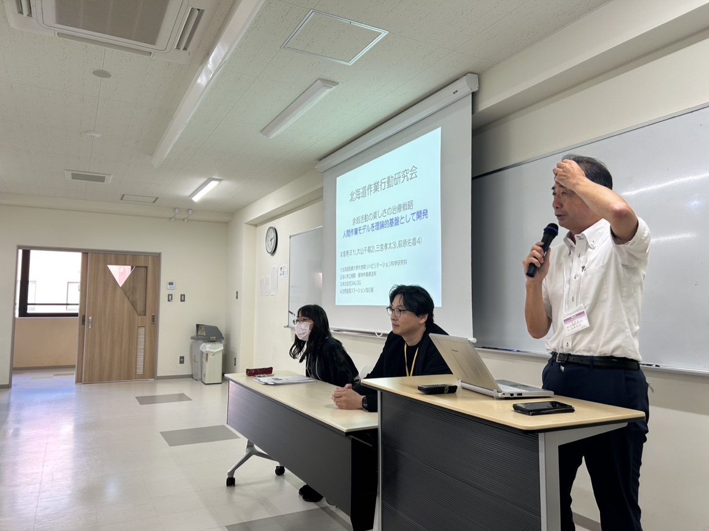 北海道作業行動研究会ワークショップ登壇の様子。スクリーンに『余暇活動の楽しさの治療戦略 ── 人間作業モデルを理論的基盤として開発』のスライドが投影されている